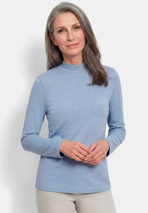Reife Frau mit langen grauen Haaren, die ein hellblaues Langarmhemd und beige Hose trägt und vor einem schlichten weißen Hintergrund steht.