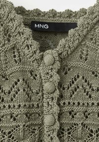 Gros plan d’un cardigan vert tricoté avec des boutons ronds texturés et un bord festonné, avec une étiquette noire portant le texte « MNG ».