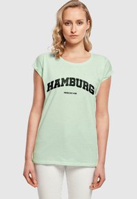Lys mintgrønn t-skjorte med korte ermer, med fet svart "HAMBURG" tekst og "MERCHC+DE" under i en enkel skrifttype.