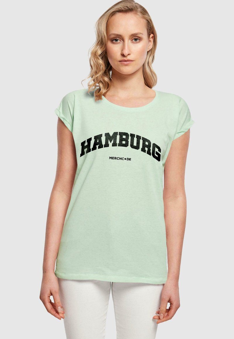 Hele piparmündiroheline T-särk lühikeste varrukate ja julge musta "HAMBURG" tekstiga, mille all on lihtsas fondis "MERCHC+DE".
