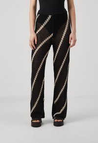 Pantalons en filet noirs avec des rayures diagonales cr�ème, design à jambes larges, taille élastique et motif texturé. Portés avec des sandales noires à semelle épaisse.
