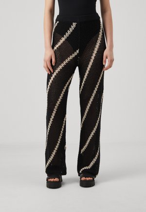 Pantalones de malla negros con rayas diagonales cremosas, diseño de pierna ancha, cintura elástica y patrón texturizado. Combinados con sandalias negras gruesas.
