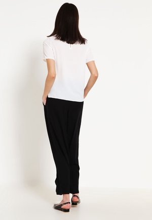 Kobieta stojąca z rękami w kieszeniach czarnych spodni, ubrana w białą bluzkę z krótkim rękawem i czarne buty z odkrytymi piętami na białym tle.