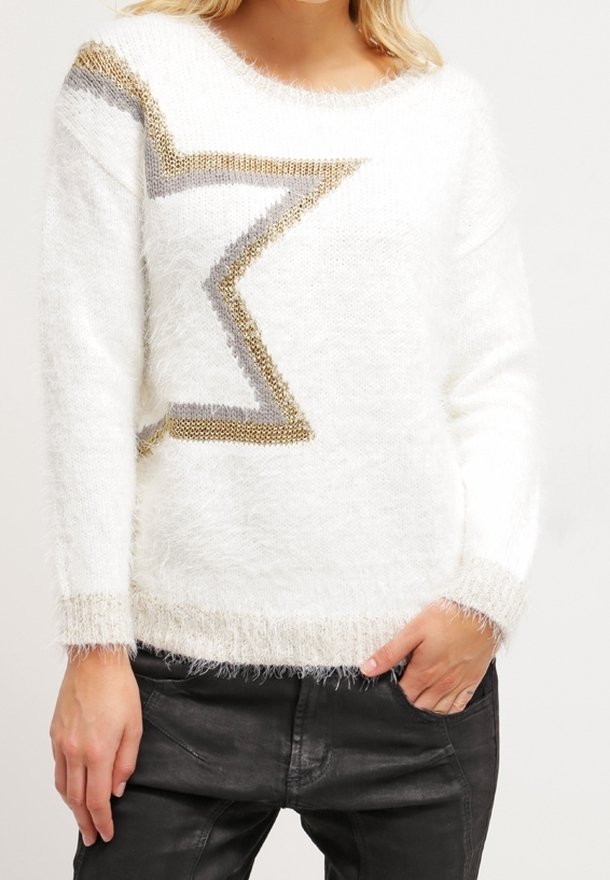 Pull blanc duveteux avec motif zigzag doré et gris, porté avec un pantalon noir ; une main dans la poche, fond neutre.