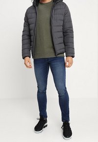Grauer Pufferjacke mit Kapuze, gestepptes Design, warmes Material. Olivgrüner Pullover darunter, blaue Slim-Fit-Jeans, schwarze Sportschuhe.