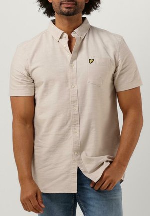 Mann trägt ein beige-farbenes kurzärmeliges Hemd mit Brusttasche und kleinem gelbem Logo, kombiniert mit blauen Jeans, vor einfachem Hintergrund.