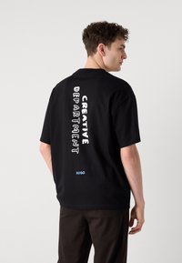 Black cotton t-shirt featuring “CREATIVE DEPARTMENT” text in white and “HUGO” in blue, short sleeves, and a relaxed fit.