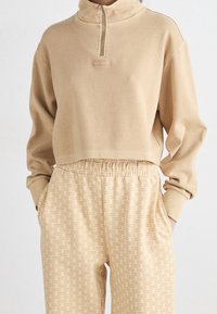 Beige cropped sweatshirt met een hoge kraag en een ritssluiting. Gecombineerd met bijpassende beige sweatpants met een logo patroon over het geheel.
