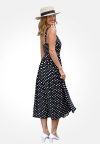 Czarna sukienka w kropki z dopasowanym górnym odcinkiem, regulowanymi ramiączkami i zwiewnym dołem; zestawiona z beżowym kapeluszem ze słomy i białymi butami na słupku.