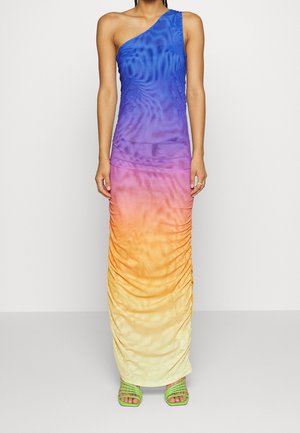 Kvinde iført en asymmetrisk maxikjole med gradient i blå, lilla, orange og gul samt grønne remsandaler, stående mod en ensfarvet baggrund.