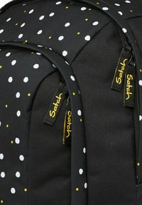 Schwarzer Rucksack mit weißen und gelben Punkten. Verfügt über robuste Reißverschlüsse mit gelber Markenkennzeichnung. Strukturierter Stoff für Langlebigkeit.