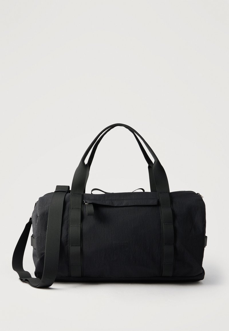 Μαύρη ορθογώνια τσάντα duffel με δύο επάνω λαβές και ρυθμιζόμενο ιμάντα ώμου, μπροστά σε απλό φόντο.