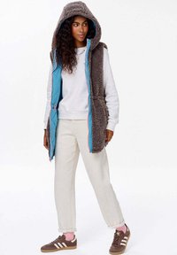 Hnědá chlupatá vesta s modrou podšívkou a růžovými akcenty, s kapucí, zipovým zapínáním a předními kapsami, nošená přes bílou mikinu.