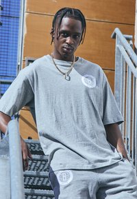 Camiseta de algodón gris de corte oversized con un detalle de logo redondo y pantalones de chándal a juego. Lleva joyería de cadena dorada frente a un fondo texturizado.