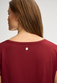 Bordeauxfarbendes Shirt mit weitem Ausschnitt und kurzen Ärmeln. Auffälliges silbernes Knopfdetail auf der Rückseite. Weicher Stoff mit glatter Textur.