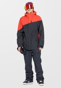 Sneeuwjack met een rode bovenhelft en een zwarte onderhelft, gemaakt van synthetisch materiaal, uitgerust met een ritszak en een verstelbare capuchon.