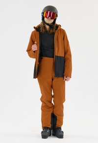 Oranje waterdichte skijas met zwarte accenten, gecombineerd met oranje skibroek. Zwarte helm en spiegeldiepte goggles. Gebruiker in een staande pose.