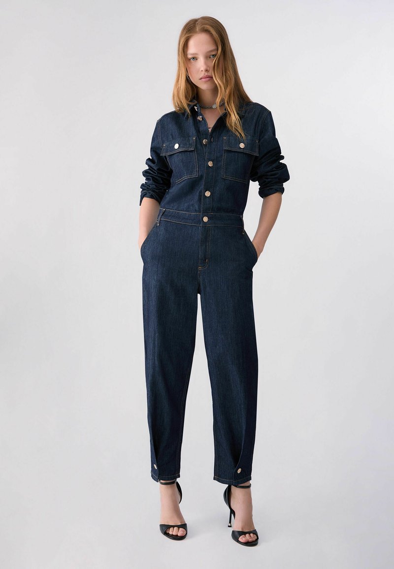 Femme aux longs cheveux portant une combinaison en denim foncé avec des boutons et des poches, debout les mains dans les poches, portant des sandales noires à talons hauts.