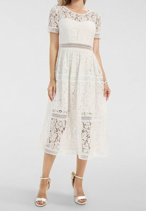 Femme portant une robe midi en dentelle blanche à manches courtes, taille cintrée et motifs floraux, associée à des sandales blanches à talons hauts.