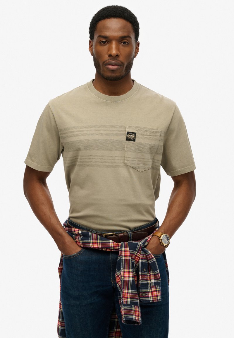 T-shirt in cotone beige con righe orizzontali e taschino frontale, abbinato a jeans blu scuro e camicia a quadri annodata in vita.