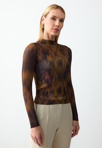 Langarmtop in transparant, geplooid stof met een golvend bruin en gouden patroon. Hoog gesloten halsontwerp, gecombineerd met lichtgekleurde broek.