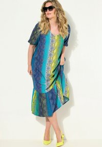 Kleid in blauem und grünem Schlangenmusterdruck, mit kurzen Ärmeln und einem gestuften Saum. Kombiniert mit gelben spitzen High Heels und großen schwarzen Sonnenbrillen.