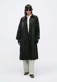 Femeie purtând o haină lungă neagră, mănuși negre, ochelari de soare negri supradimensionați, cămașă albă, pantaloni deschis la culoare și pantofi maro, stând în fața unui fundal alb.