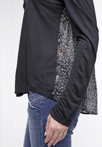 Zwarte langemouwen top met een doorzichtige bloemenpaneel aan de zijkant. Beschikt over een ronde zoom en knopen aan de onderkant. gedragen met blauwe jeans.