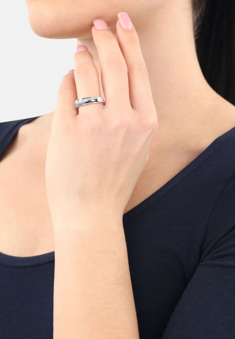 Hand med polerade naglar som bär en silverring med små diamanter, person i marinblå topp mot en vit bakgrund.