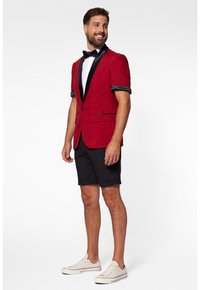 Crveni sako s cvjetnim uzorkom i crnim reverima, uparен s crnim šorcima i bijelim tenisicama. Leptir mašna naglašava formalni izgled.