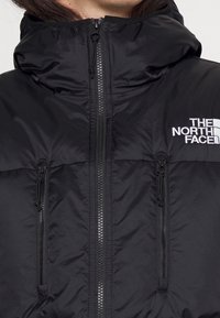 Sort dunjakke med hætte, to lynlåslommer i siden og hvid logo-broderi på højre bryst. Glat, blank tekstur.