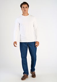 Pitkähihainen valkoinen puuvillapaita, joka on yhdistetty sinisiin farkkuihin ja ruskeisiin veneen kenkiin. Paita on varustettu nauhakiinnityksellä ja siinä on rento leikkaus.