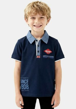 Garçon blond souriant portant un polo bleu marine avec un col bleu clair et un détail du drapeau britannique, debout devant un fond blanc.