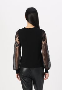 Pull noir avec manches longues en dentelle transparente ornées de motifs floraux. Le corps est de couleur noire unie, et les poignets sont côtelés. Porté avec un pantalon noir.