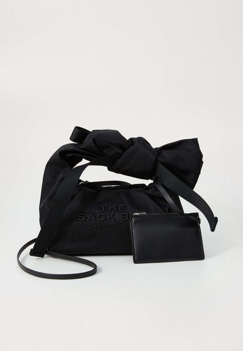 Bolso de nylon negro con parte superior anudada y texto en relieve, acompañado de un tarjetero de cuero negro. Forma plana, diseño elegante, acentos mínimos.