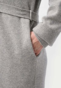 Grå stickad plagg med midjeband, som visar en texturerad tyg och sidofickor; mjukt utseende och finish med ribbad ärmslut.