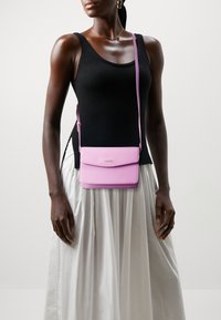 Růžová crossbody taška s texturovaným povrchem a klopou, zdobená decentním logem a nastavitelným popruhem. Nositá s černým tílkem.