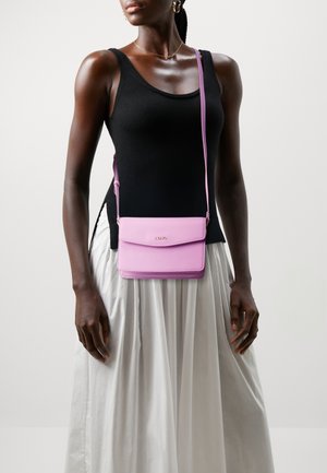 Růžová crossbody taška s texturovaným povrchem a klopou, zdobená decentním logem a nastavitelným popruhem. Nositá s černým tílkem.