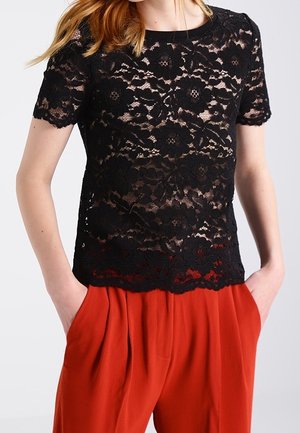 Nainen, joka käyttää mustaa kukallista pitsistä lyhythihaista yläosaa nude-aluskerroksen päällä ja löysiä punaisia korkeavyötäröisiä housuja, kädet taskuissa.