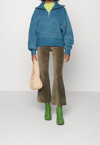 Pull-over en maille bleue avec col à zip, associé à un pantalon évasé en velours côtelé olive, des bottines vertes et un sac à main texturé beige.