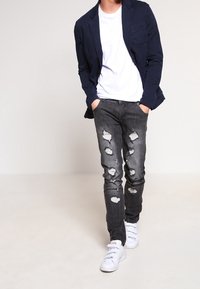 Blazer blu navy sopra una maglietta bianca, abbinato a jeans neri strappati e sneakers bianche. I jeans presentano evidenti toppe rovinate.