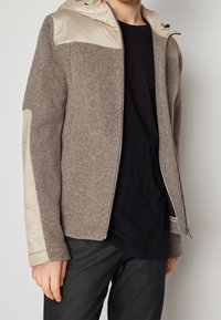 Giacca grigia in misto lana con cappuccio in nylon beige, chiusura con zip e pannelli laterali. Presenta una finitura strutturata e un design aderente.