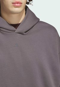 Dugulja s kapuljačom u sivo-ljubičastoj tkanini s mekom teksturom. Ima mali otisnuti logo na području prsiju.