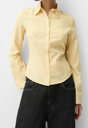 Femme portant une chemise ajustée jaune à manches longues avec des rayures blanches verticales et un pantalon taille haute foncé.