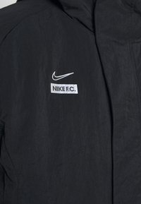 Veste noire avec logo Nike swoosh blanc et texte "NIKE F.C." sur la poitrine, montrant une partie de la zone de la fermeture éclair avant.