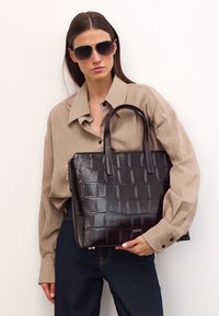 Tummanruskea krokotiili-kuosiselkäinen olkalaukku, jossa on kaksi kahvaa, kiiltävä pinta ja vetoketjukiinnitys. Yhdistetty beigen raidallisen paidan ja tummien farkkujen kanssa.