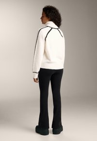 Maglione bianco morbido con cuciture nere a contrasto, collo alto. Abbinato a pantaloni neri svasati e scarpe platform verde scuro. Design minimalista.