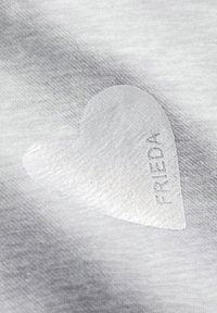 Grauer Stoff mit einer glatten Textur weist einen weißen, herzförmigen Patch auf, der mit dem Wort "FRIEDA" in subtiler metallischer Schrift geprägt ist.