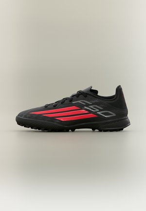 Chaussure de sport noire avec des rayures rouges et des lacets noirs, conçue pour les surfaces en gazon synthétique, présentée en profil latéral sur un fond neutre.