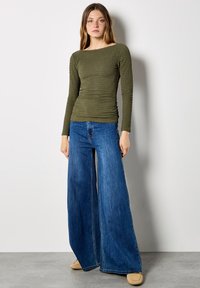 Olive green, long-sleeve, fitted top with textured, ruched fabric paired with high-waisted, wide-leg, light blue jeans. Smooth, soft shoes.